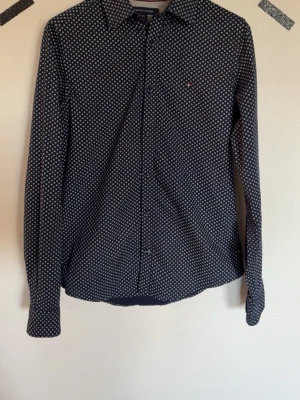 Mönstrad skjorta från Tommy Hilfiger - Snygg mörkblå skjorta från Tommy Hilfiger med vitt prickigt mönster. Klassisk krage, långa ärmar och knäppning framtill. Liten logga på bröstet. Perfekt för dig som gillar stilrena detaljer och bekvämt material.