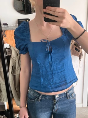 Blå linnetopp med puffärm och knyt - Superfin blå topp i linne med puffärmar och fyrkantig urringning. Toppen har en söt knytning framtill och är croppad i modellen. Perfekt att styla med jeans eller kjol för en trendig look.