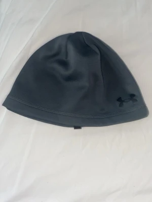Mörkgrå mössa från Under Armour - Snygg och stilren mörkgrå mössa från Under Armour med diskret logga framtill. Tillverkad i ett mjukt och stretchigt funktionsmaterial som passar perfekt för kyliga dagar. Enkel design som funkar till det mesta.