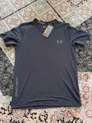  Under Armour t shirt - Svart kortärmad träningströja från Under Armour med logga på bröstet och tryckt text längs sidan. Tillverkad i ett lätt och snabbtorkande funktionsmaterial som passar perfekt för träning och sportaktiviteter. Modern passform och rund hals.