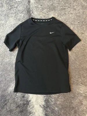 Svart Nike träningstopp - Snygg svart träningstopp från Nike med klassisk logga på bröstet. Tillverkad i ett lätt och funktionellt syntetmaterial som andas. T-shirten har rund halsringning och korta ärmar, perfekt för aktiva dagar.