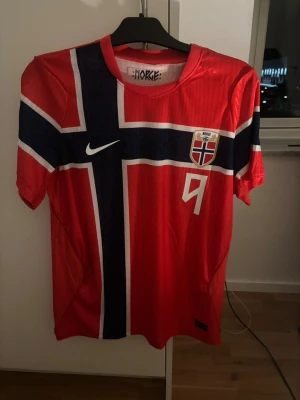 Norge fotbollströja Nike Haaland #9 - Röd Norge fotbollströja från Nike med stort kors i blått och vitt, officiellt landslagsmärke och nummer 9 på bröstet. På ryggen står det 'Braut Haaland' och siffran 9. Perfekt för dig som vill stötta Norge på match eller träning.