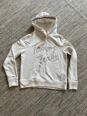 Vit hoodie från Tommy Jeans - Snygg vit hoodie från Tommy Jeans med stor logga framtill och klassisk huva med dragsko. Tröjan har ribbade muddar vid ärmslut och nederkant samt diskreta detaljer på ärmarna. Perfekt för en avslappnad och trendig look. Str S. Två små fläckar(se bild)