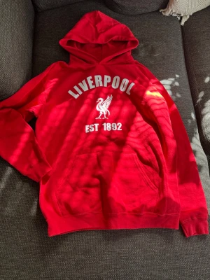 Röd Liverpool hoodie med tryck - Helt nytt skick, aldrig använd. För liten för mig och lillebror som håller på annat lag. Väldigt snygg röd färg och perfekt för dig som hejar på Liverpool. Köpt i butik.