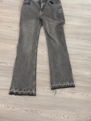 Grå Zara flared jeans - Säljer ett par grå jeans med rak passform och råa, fransiga benslut. Jeansen har klassisk femficksmodell och hög midja. Perfekta för en avslappnad och trendig look.