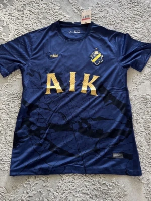 Blå AIK matchtröja från Nike - Snygg blå AIK matchtröja från Nike med klubbmärke broderat på bröstet och stora gula bokstäver över magen. Tröjan har rund hals och är tillverkad i ett lätt, ventilerande material perfekt för träning eller match.