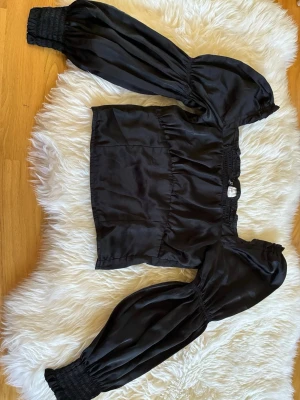 HM Svart satintopp med puffärmar - Snygg svart topp i satin med långa puffärmar och smock vid ärmslut och axlar. Toppen har en croppad passform och en lätt glansig yta som ger en lyxig känsla. Perfekt att styla med höga jeans eller kjol.