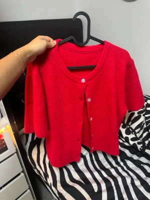Röd stickad kofta med kort ärm - Snygg röd stickad kofta med korta ärmar och rund halsringning. Koftan har knäppning framtill med vita knappar och är perfekt för att ge din outfit en färgklick. Enkel och stilren design som passar till många looks.