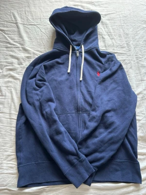 Marinblå hoodie från Polo Ralph Lauren - Snygg marinblå hoodie från Polo Ralph Lauren med dragkedja, vit dragsko och den klassiska röda loggan broderad på bröstet. Tillverkad i mjuk bomullsblandning och har en stor huva för extra komfort. Perfekt för en avslappnad och stilren look.
