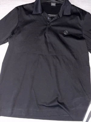 Svart pikétröja från BOSS - Klassisk svart pikétröja från BOSS med korta ärmar och krage. Tröjan har knappar framtill och en diskret logotyp på bröstet. Perfekt för en stilren och avslappnad look.