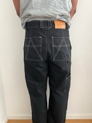 Svarta jeans från Axel Arigato - Svarta jeans från Axel Arigato med raka ben, kontrasterande vita sömmar och stora bakfickor med unikt mönster. Jeansen har en brun läderpatch med logga i midjan och breda hällor. Perfekta för en trendig streetwear-look.