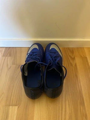 Blå Nike CR7 fotbollsskor - Säljer ett par blå Nike mercurial  veloce CR7 fotbollsskor med vita detaljer och snörning. Skorna har en smal passform och är designade för spel på gräs. Snygg CR7-logga på hälen och klassisk Nike-swoosh på ovansidan. Perfekta för dig som vill sticka ut på planen.