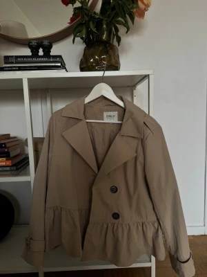 Beige trenchcoat från ONLY - Stilren beige trenchcoat från ONLY med dubbelknäppning framtill, bred krage och volangdetalj nedtill. Jackan har långa ärmar med justerbara slejfar vid handlederna och är tillverkad i ett lätt material som passar perfekt till vår och höst. Andvänd fåtal gånger 