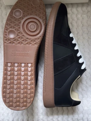 Svarta Maison Marigelas  - Stilrena svarta sneakers från Maison Margiela med vita skosnören och klassisk brun gummisula. Ovandelen är i en mix av läder och mocka, vilket ger en snygg kontrast. Diskret logotyp på sulan och ikonisk design inspirerad av tyska arméskor.             Vid några funderingar är det bara att höra av sig☺️