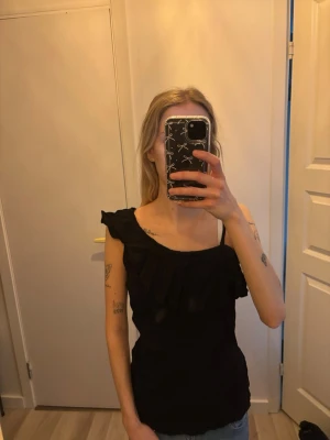  One shoulder topp med volang - Snygg svart oneshoulder topp med volangdetalj längs halsringningen. 