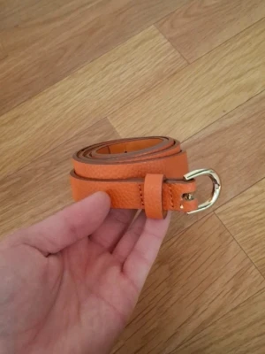 Smal orange skinnbälte med gulddetalj - Snyggt smalt bälte i orange skinn med guldfärgat spänne. Perfekt för att ge din outfit en färgklick och passar till både jeans och klänning. Bältet har en stilren design och flera hål för justerbar passform. Pyttelite avskavt,syns inte när det är knäppt 