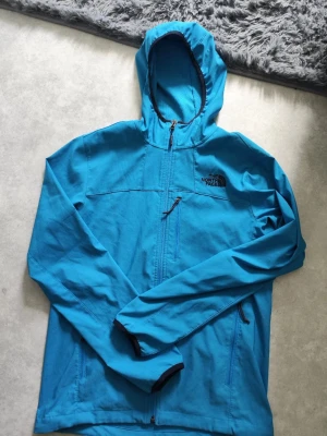 Blå vindjacka från The North Face - Snygg blå vindjacka från The North Face med huva och dragkedja framtill. Jackan har en bröstficka med dragkedja och logga på vänster sida. Perfekt för blåsiga dagar och outdoor-aktiviteter. Lätt material och sportig look.