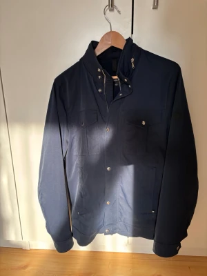 J.lindeberg fieldjacket - Snygg mörkblå jacka med ståkrage och tryckknappar. Två bröstfickor med lock och knapp, samt diskreta sidofickor. Jackan har en minimalistisk design och är tillverkad i ett slätt tyg som ger en stilren look. Perfekt för dig som gillar enkel men trendig stil.