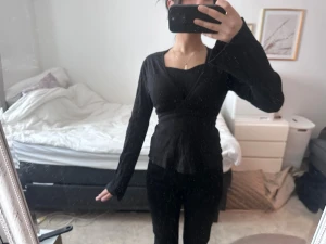 Svart omlottblus med långa ärmar - Snygg svart omlottblus med långa ärmar och markerad midja som knyts i ryggen. Blusen har en fyrkantig ringning och sitter figurnära, perfekt för en trendig och stilren look.