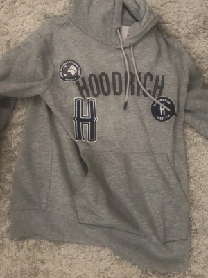 Grå hoodie från Hoodrich med tryck - Snygg grå hoodie från Hoodrich med stora tryck och patches på bröstet. Tröjan har huva med dragsko och en stor ficka framtill. Materialet är mjukt och skönt, perfekt för en avslappnad streetwear-look.