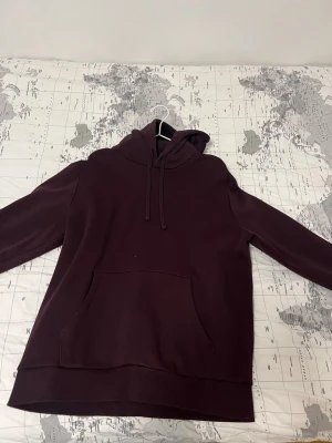 Vinröd hoodie med magficka - En stilren vinröd hoodie med huva och dragsnören. Hoodien har en stor magficka framtill och är tillverkad i ett mjukt och bekvämt material. Perfekt för en avslappnad och trendig look.