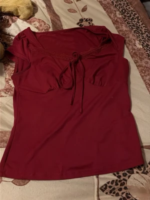 Vinröd topp med knyt och spets - Snygg vinröd topp med knyt framtill och spetsdetalj vid urringningen. Modellen är ärmlös och har en feminin passform. Perfekt att styla med jeans eller kjol.
