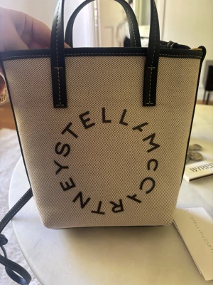 Stella McCartney beige handväska - Trendig beige handväska från Stella McCartney med svart text i cirkelformad logga på framsidan. Väskan har svarta handtag och detaljer samt axelrem. Tillverkad i canvasliknande material med kontrastsömmar. Har även avtagbara axelremmar så den kan bäras crossbody. Slutsåld på en halvtimme. 