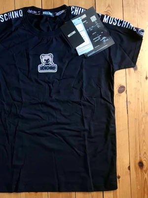 Svart Moschino t-shirt med björnlogga - Svart t-shirt från Moschino med korta ärmar och rund hals. På bröstet finns en broderad björnlogga och Moschino-tryck längs axlarna. Tillverkad i mjuk bomull, perfekt för en trendig och avslappnad stil.