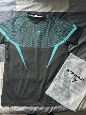 Svart och turkos träningströja Gymshark - Snygg svart träningströja från Gymshark med turkosa detaljer och logga på bröstet. Tröjan har rund hals och är tillverkad i ett lätt och stretchigt material som andas, perfekt för gym eller löpning.