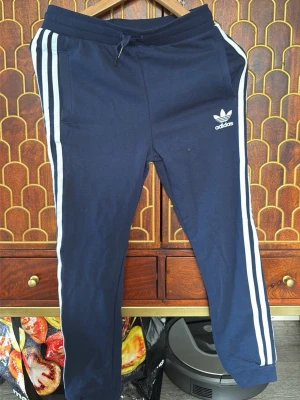 Marinblå Adidas mjukisbyxor - Mörkblå mjukisbyxor från Adidas med klassiska vita ränder längs sidorna och broderad logga på vänster ben. Byxorna har dragsko i midjan och sidofickor. Perfekta för en avslappnad och sportig stil.