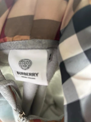 Grå zip hoodie från Burberry - Snygg grå zip hoodie från Burberry med klassiskt rutigt foder i huvan och diskret broderad logga på bröstet. Tillverkad i mjuk bomullsblandning och har dragsko i huvan. Perfekt för en chill och stilren look. Det står M men det är S