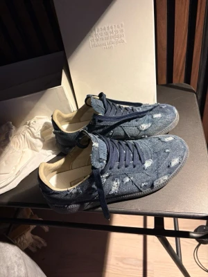 Maison Margiela denim sneakers - Snygga sneakers från Maison Margiela i blått denimtyg med distressed detaljer och mörkblå skosnören. Skorna har en klassisk låg siluett och beige innerfoder. Perfekta för dig som gillar unik streetstyle och designer vibes.