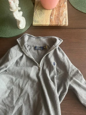 Grå halvzip från Polo Ralph Lauren - Snygg grå långärmad tröja från Polo Ralph Lauren med halv dragkedja och klassisk broderad logga på bröstet. Tröjan har ribbade muddar och krage, perfekt för en stilren och avslappnad look.
