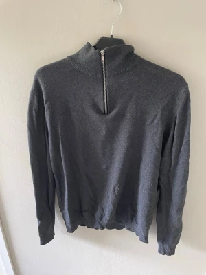 Mörkgrå tröja från Jack&Jones med halfzip  - Mörkgrå långärmad tröja med hög krage och halv dragkedja framtill. Tröjan har ribbade muddar vid ärmslut och nederkant. Perfekt för lager-på-lager och enkel att matcha med jeans eller chinos.