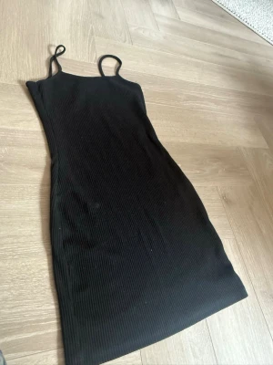 Svart ribbad kort klänning - Svart ribbad klänning med smala axelband och kroppsnära passform. Klänningen är ärmlös och har en enkel, stilren design som passar perfekt till många olika tillfällen.