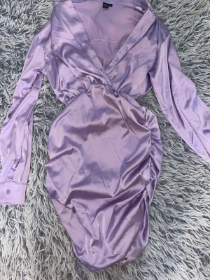 Ljuslila omlottklänning i satin - Snygg ljuslila omlottklänning i glansigt satinmaterial med v-ringning och långa ärmar. Klänningen har draperade detaljer längs sidorna och en figurnära passform som ger en elegant siluett. Perfekt för dig som vill sticka ut med en trendig look.