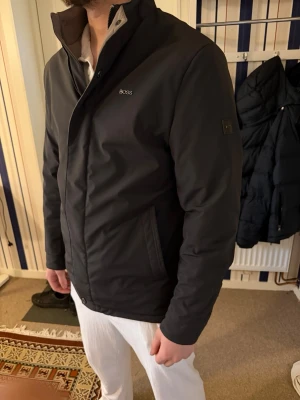 Svart jacka från BOSS med dragkedja - Snygg svart jacka från BOSS med diskret logga på bröstet och ärmen. Jackan har hög krage, dragkedja och knappar framtill samt två sidofickor. Perfekt för dig som vill ha en stilren och modern look. Nypris:5900