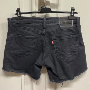 Svarta jeansshorts från Levi's - Modell 511, storlek w33 l32. Svarta jeansshorts från Levi's med råa, fransiga benslut och klassiska femficksdetaljer. Shortsen har normal passform och midwaist, med Levi's ikoniska läderpatch bak och röd logotag på bakfickan. Perfekta för en chill sommarstil.