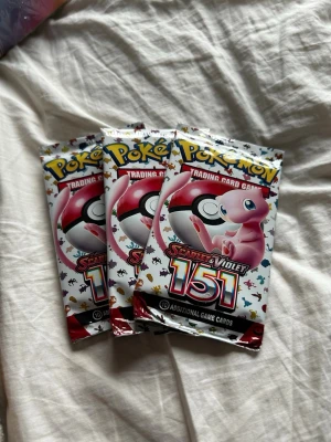 Pokémon Scarlet & Violet 151 boosterpack - Tre oöppnade Pokémon Scarlet & Violet 151 boosterpack med Mew på framsidan. Varje paket innehåller 10 samlarkort. Perfekt för dig som vill utöka din samling eller jaga efter sällsynta kort. Förpackningarna är färgglada med vit bas och röda detaljer.3 packs oöppnade
