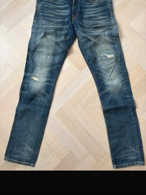 Blå slitna jeans med raka ben - Säljer ett par blå jeans med raka ben och slitna detaljer på både fram- och baksida. Jeansen har klassisk femficksdesign och tydliga slitningar och skrapmärken för en cool, avslappnad look. Perfekta för dig som gillar en streetstyle-vibe.