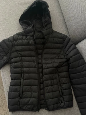 Svart pufferjacka med huva - Säljer en svart pufferjacka med huva och dragkedja framtill. Jackan har två fickor med dragkedja och quiltad design. Perfekt för kalla dagar och ger en sportig look. Materialet är lätt och isolerande.