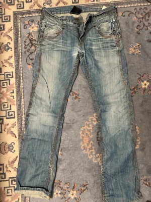 Ljusblå jeans med kontrastsömmar - Säljer ett par ljusblå jeans med snygga slitningar och kontrastsömmar. Jeansen har fem fickor och coola detaljer på bakfickorna. Klassisk rak passform och tillverkade i denim. Perfekta för en avslappnad stil.