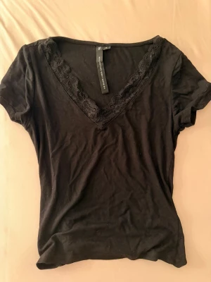 Svart v-ringad topp med spets - Svart kortärmad topp från The Black Label Collection med v-ringning och dekorativ spetskant runt halsen. Tunn och mjuk i materialet, perfekt att styla med jeans eller kjol. Enkel men ändå med en snygg detalj i spetsen. Storlek S använd 3 gånger max