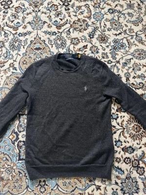 Mörkgrå merinoulltröja Polo Ralph Lauren - Mörkgrå slim fit tröja från Polo Ralph Lauren i mjuk och tvättbar merinoull. Klassisk rund halsringning och diskret broderad logga på bröstet. Perfekt för en stilren och bekväm look.  Storlek M barn eller 164cm