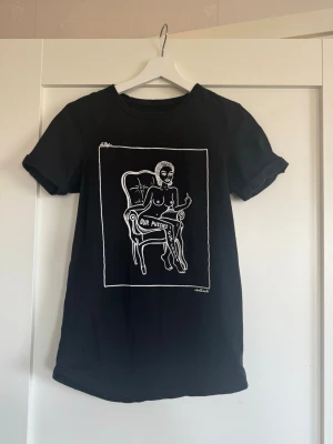 Svart t-shirt med konsttryck  - Svart t-shirt skapad av lokala konstnärer i Amsterdam, med  vitt konsttryck framtill som föreställer en person i en stol och texten 'OUR PUSSIES, OUR CHOICES.'. T-shirten har korta ärmar och rund hals. Perfekt för dig som gillar statement-plagg och konstnärlig stil. Står XXS men passar S.