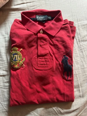 Röd pikétröja från Ralph Lauren - Röd pikétröja från Ralph Lauren med klassisk krage, knappar framtill och broderad logga på bröstet. Tröjan har korta ärmar och ett broderat emblem på vänster sida. Perfekt för en sportig och stilren look.