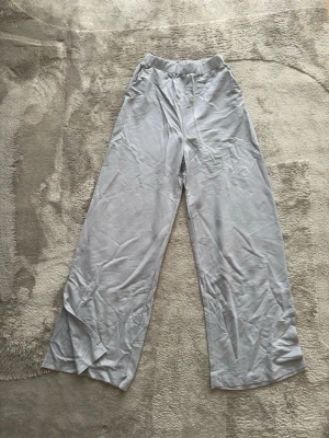 Vida ljusgrå tygbyxor med resår - Säljer ett par ljusgrå tygbyxot med bred resår i midjan och vida ben. Materialet är mjukt och stretchigt, perfekt för en avslappnad och trendig look. Passar dig som gillar bekväma och stilrena byxor.