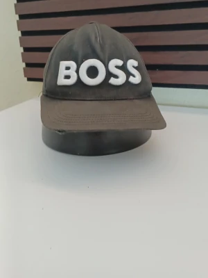 Svart keps från BOSS med brodyr - Snygg svart keps från BOSS med stor vit broderad text framtill. Klassisk böjd skärm och justerbart spänne bak för perfekt passform. Tillverkad i bomullstyg som andas. Perfekt accessoar för en cool och avslappnad stil.