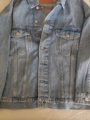 Klassisk ljusblå jeansjacka - Snygg ljusblå jeansjacka med raka linjer, klassisk krage och silverfärgade knappar. Två bröstfickor med lock och knapp samt sömdetaljer framtill. Perfekt att slänga över en hoodie eller t-shirt för en avslappnad look.