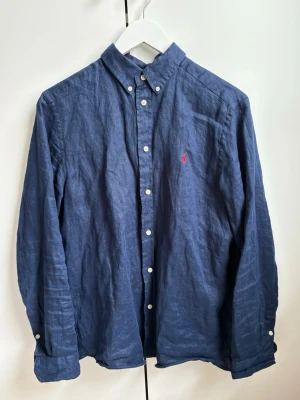 Blå linneskjorta från Ralph Lauren - Snygg blå skjorta i linne från Ralph Lauren med klassisk button-down krage och vita knappar. 